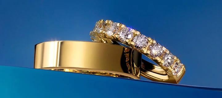 Dwie złote obrączki na niebieskim tle: gładka szeroka obrączka i ozdobna z rzędowymi diamentami.