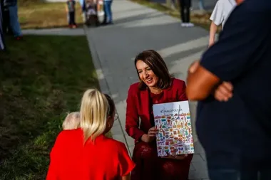 Kobieta w czerwonym garniturze klęczy na chodniku i wręcza torbę z napisem „Małopolska” kobiecie trzymającej dziecko, podczas spotkania na zewnątrz.